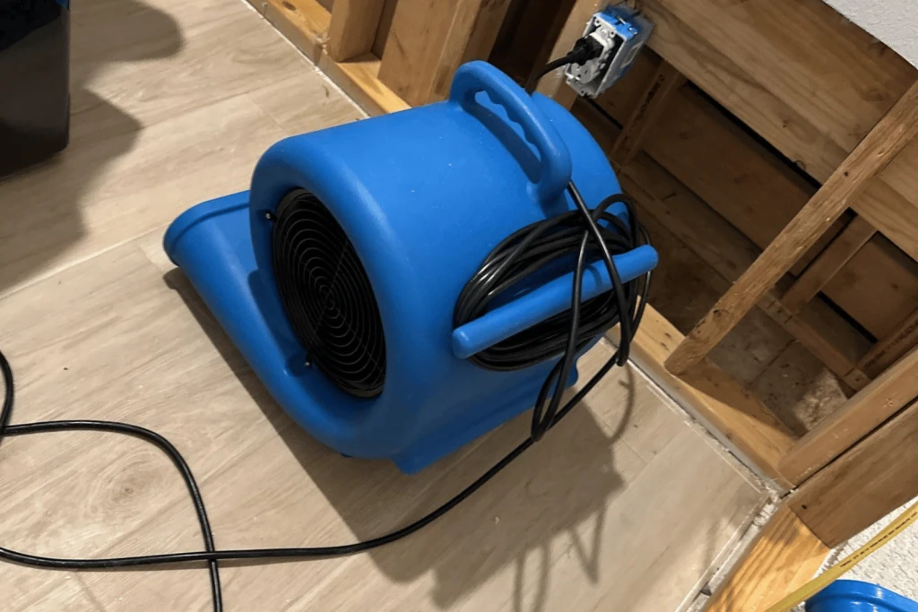 Blue air mover on a floor beside exposed framing.