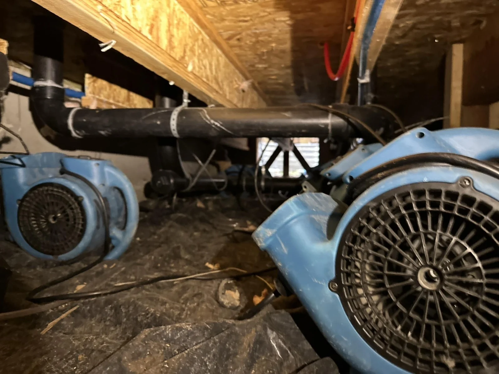 Blue air movers positioned beneath exposed floor joists and plumbing in a crawl space