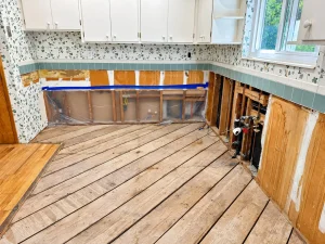 Kitchen after water damage with lower cabinets removed, drywall flood-cut to expose framing and plumbing, and plastic containment over the open wall